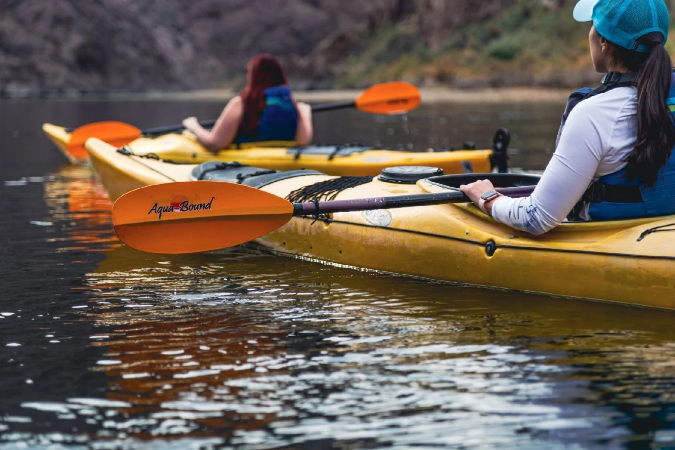 Two people kayaking on calm water with yellow kayaks and orange paddles.